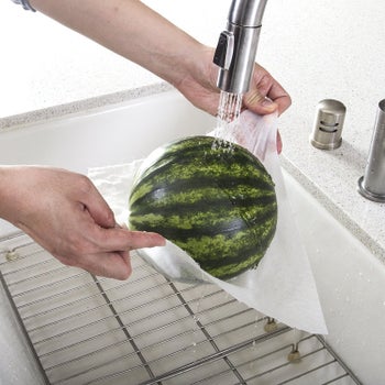 Hands washing a watermelon in a sink, holding it in the  towel, which isn't ripping under the weight