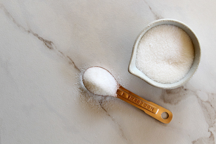 Measuring cup and spoon full of granulated sugar on top of a marble countertop.