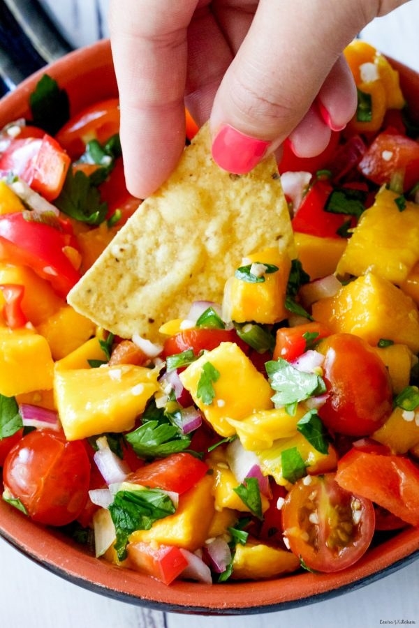 A hand is dipping a tortilla chip into a bowl of fresh salsa made with chunks of mango, cherry tomatoes, onions, and cilantro