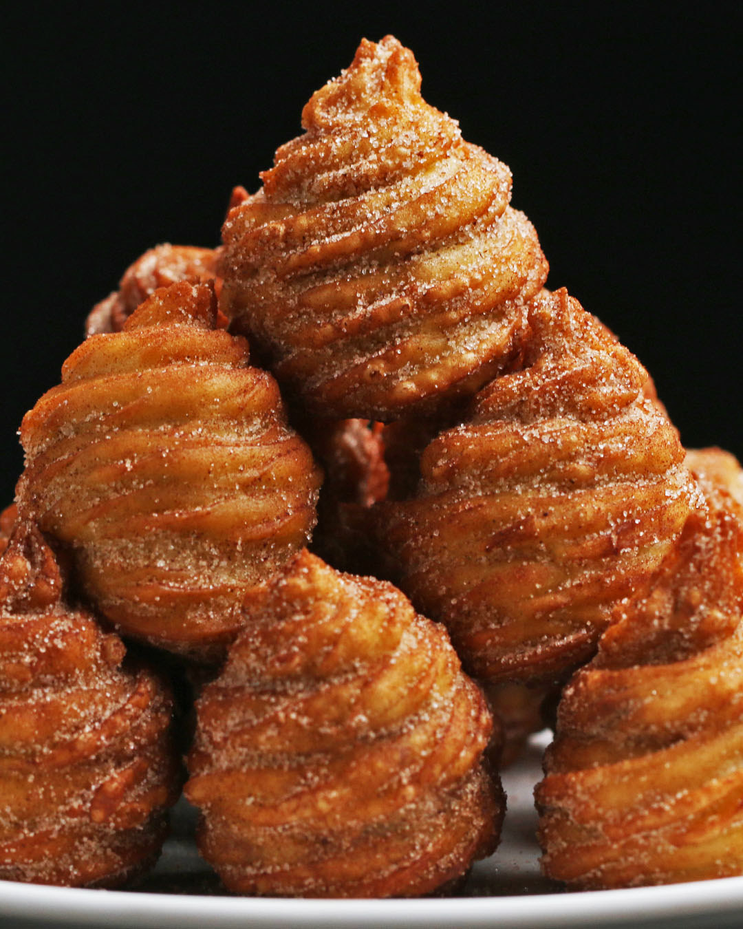 Bombas de churrosRende 18 bombas de churrosVocê vai precisar de: 1 xícara de água4 colheres de sopa (½ barra) de manteiga½ xícara e 2 colheres de sopa de açúcar (separados)½ colher de chá de sal1 xícara de farinha de trigo tipo 13 ovos grandes1 colher de chá de extrato de baunilha18 trufas de chocolate ao leiteÓleo, para fritar½ xícara de açúcar2 colheres de sopa de canela1 saco de confeitar com um bico fechado pequeno em formato de estrelaModo de preparo: 1. Em uma panela média em fogo médio-alto, misture a água, a manteiga, 2 colheres de sopa de açúcar e o sal e deixe ferver.2. Quando a mistura começar a ferver, diminua a temperatura para médio-baixo e acrescente a farinha. Usando uma colher de madeira, mexa até que a massa fique homogênea e forme uma bola, por cerca de um minuto.3. Remova a panela do fogo e deixe esfriar por 5 minutos antes de continuar, de modo que os ovos não cozinhem.4. Misture os ovos, um de cada vez, incorporando totalmente cada um antes de adicionar o próximo. Em seguida, adicione a baunilha. 5. Forre uma assadeira com papel manteiga. 6. Coloque a massa em um saco de confeitar com um bico fechado em formato de estrela.7. Faça 18 círculos de 4cm sobre a folha de papel manteiga. Coloque uma trufa de chocolate ao leite em cima de cada um. Faça um espiral com a massa para cobrir as trufas, sendo cuidadoso para selar totalmente o chocolate. Transfira imediatamente a assadeira para o congelador e deixe por uma hora, até que fiquem sólidos.8. Aqueça o óleo em uma panela funda até atingir 180°C. 9. Leve ao forno de 4 e 4 até que fiquem dourados, por cerca de 3 minutos, virando de modo que fritem de maneira uniforme. Coloque sobre um prato com papel toalha e tire o excesso de óleo.10. Misture a xícara de açúcar e a canela em uma tigela pequena. Cubra a bomba de churros com a mistura.11. Bom apetite!Inspired by: https://tasty.co/recipe/churro-ice-cream-bowls https://cooking.nytimes.com/recipes/1825-churroshttps://smittenkitchen.com/2016/03/churros/https://www.kingarthurflour.com/recipes/churros-recipe