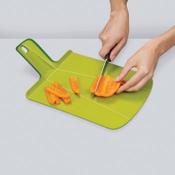 The green cutting board with handle, flat on a counter with sliced peppers on it