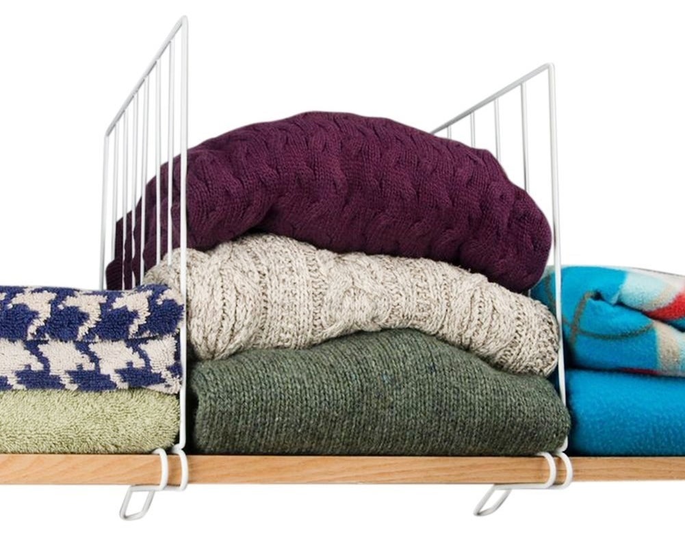 wooden shelf with white wire shelf dividers in between vertical stacks of sweaters and blankets