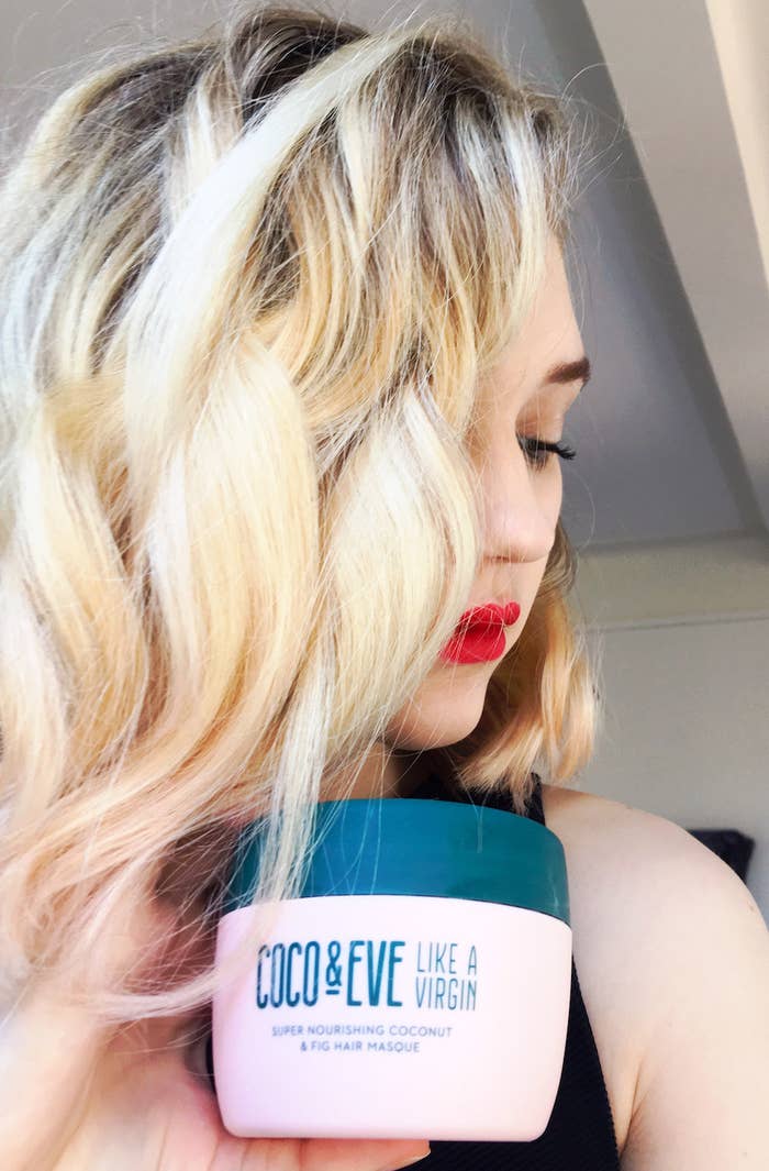 A person with wavy and shiny hair holding a hair mask container