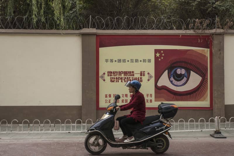 再教育施設の前をバイクで通り過ぎる女性。その塀には、プロパガンダ用ポスターが貼られている。2017年10月、カシュガル市で撮影。