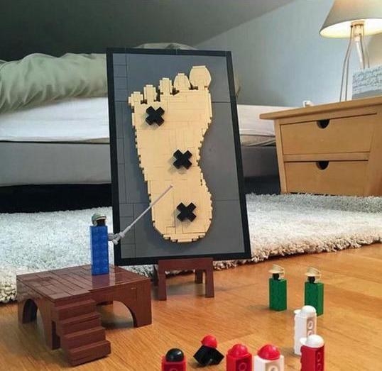 A Lego piece standing on a platform near a person&#x27;s bed pointing to a poster of a large human foot made out of Legos, with the vulnerable parts marked with an x, while other Legos stand and watch