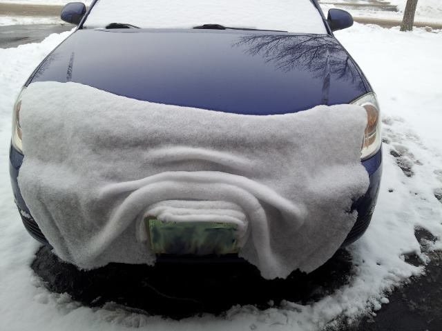 A car with a sheet of snow falling off the hood that makes it look like a blanket