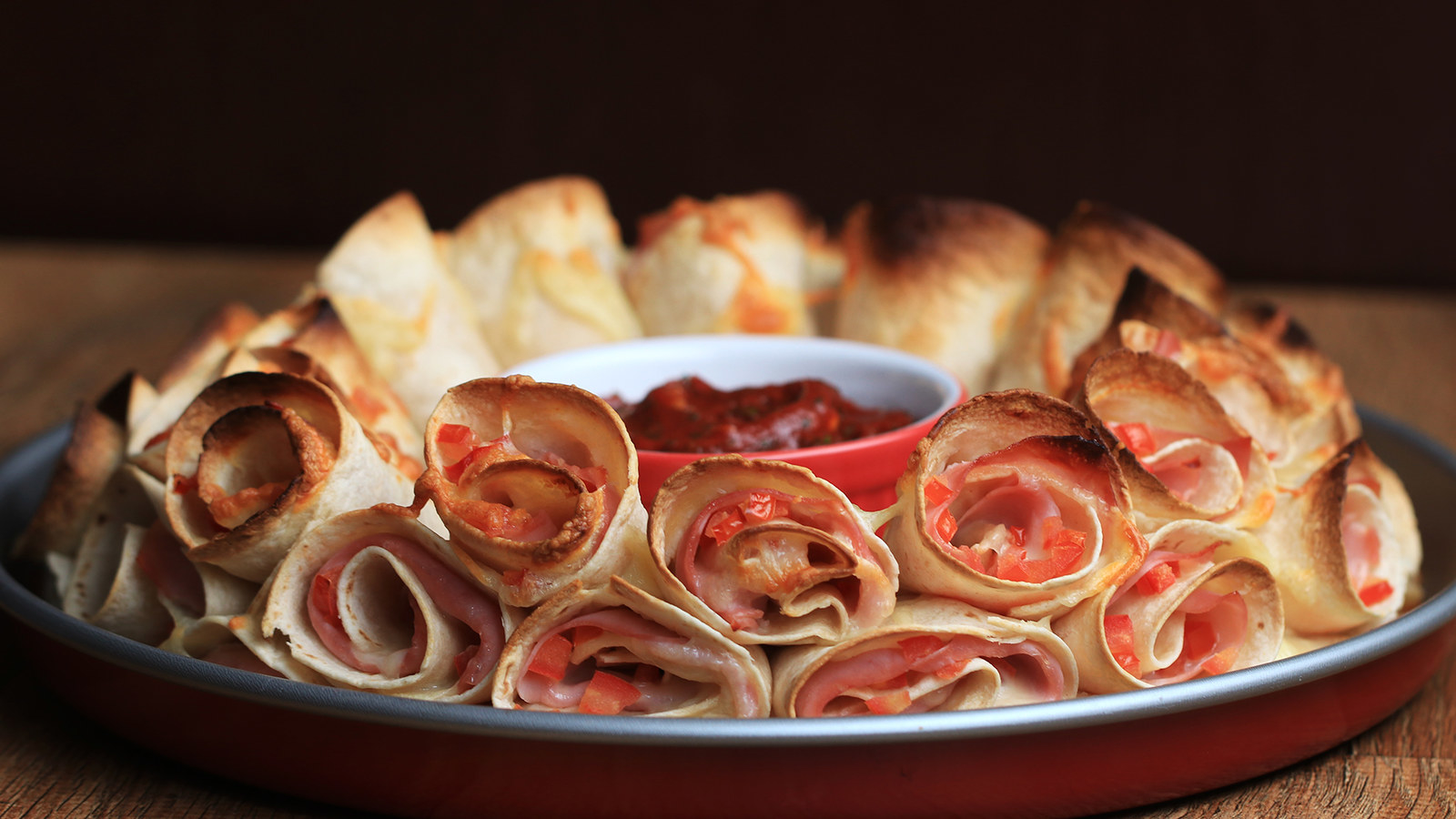 Redimento: 40 conesVocê vai precisar de:20 tortilhas de trigo200g requeijão cremoso1 xícara tomate picado20 fatias de presunto cortadas ao meio20 fatias de queijo muçarela cortadas ao meio1 xícara de queijo muçarela raladoMolho:1 xícara de extrato de tomate1 colher de sopa de orégano seco2 colheres de sopa de açúcar1 colher de chá de pimenta calabresa¾ colher de chá de sal1 colher de sopa de salsa picada1 dente de alho amassado2 colheres de sopa de azeiteModo de preparo:1. Preaqueça o forno a 180°C.2. Corte cada tortilha ao meio.3. Espalhe em cada metade o requeijão e salpique um pouco do tomate. Coloque 1/2 fatia de muçarela e ½ fatia de presunto. Repita com o restante dos ingredientes.4. Enrole cada um em formato de cone. Coloque em uma forma redonda com um pote no meio, apenas para dar estrutura. Polvilhe a muçarela e repita até obter três camadas5. Remova o pote e leve ao forno por 15-20 minutos até dourar.6. Enquanto isso, faça o molho: em uma tigela média, adicione o extrato de tomate, o orégano, o açúcar, a pimenta, o sal, a salsa, o alho e o azeite e misture. Tampe com um plástico filme e leve ao microondas por 2 minutos. 7. Retire os cones do forno e coloque e transfira o molho para uma tigela menor, que caiba no centro dos cones.8. Aproveite!