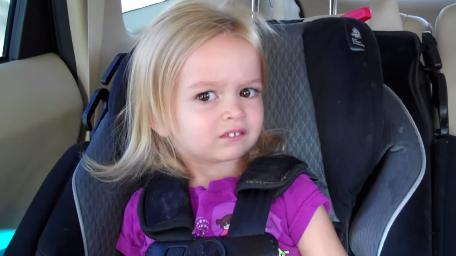 Young girl with two front teeth in a car seat looking at the camera with disgust