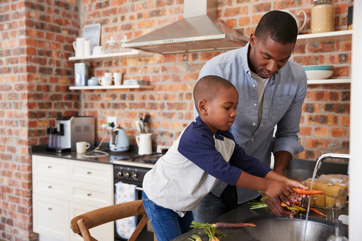 Little boy helping his dad wash carrots in a kitchen sink. Their kitchen is surrounded by exposed brick walls and shelves filled with kitchenware