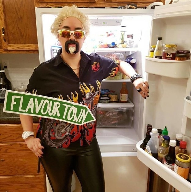 A person standing inside a fridge with a &quot;flavourtown&quot; sign. They are also wearing a wig, sunglasses, fake mustache and goatee, and a shirt with flames, 