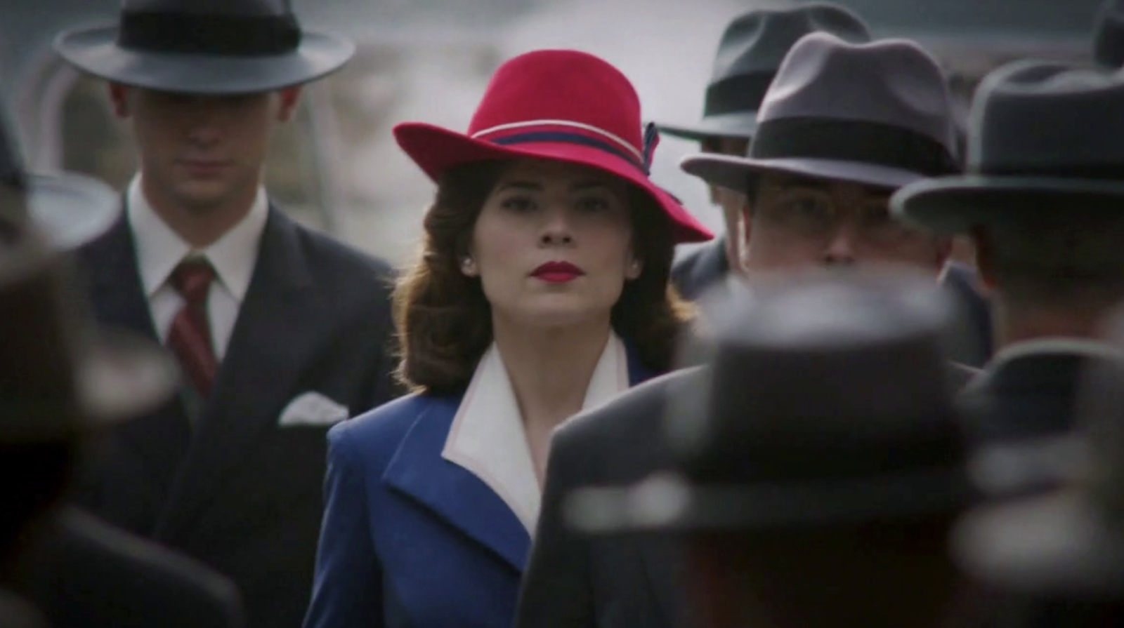 Hayley Atwell as Peggy Carter in a hat and suit amid a crowd of men in hats and suits
