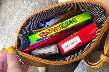 Open purse containing Sour Patch Kids candy, a chocolate bar, and other small items. Hand with painted nails holds the purse