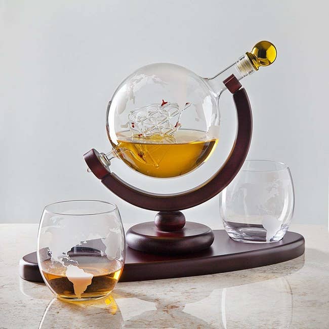 circular decanter with a glass ship in the inside and a wood base with two clear glasses with an etching of the world on them 
