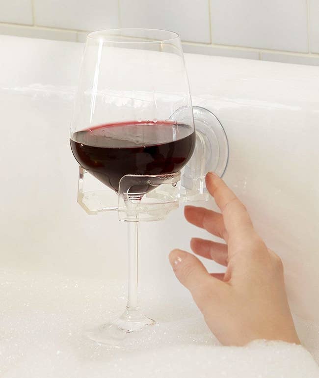 Tthe clear holder suctioned to the side of the tub with a glass of red wine in it and a hand reaching for it