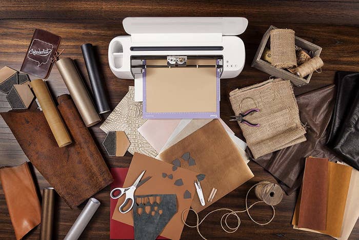 A Cricut Maker on a table covered in vinyl fabric, leather, burlap, twine, and paper goods