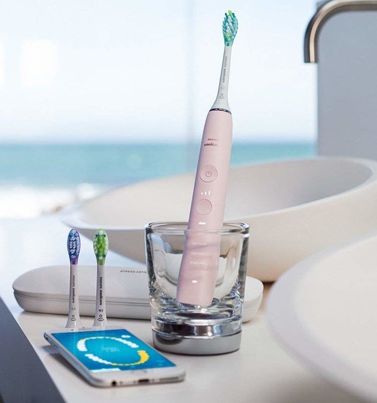 The pink toothbrush in a charging glass with replacement heads, travel case, and phone showing the app