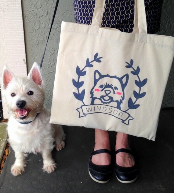 A person holding a white tote bag with a navy blue illustration of their dog and the dog&#x27;s name printed beneath the illustration