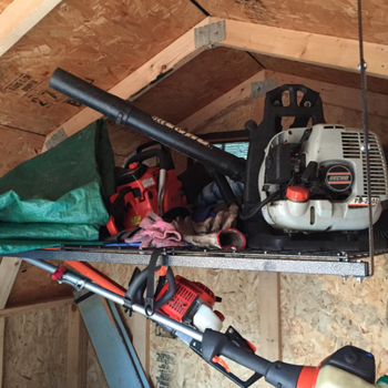 reviewer's shelf holding a leaf blower, a weed whacker, and other lawn tools near the roof of a storage shed