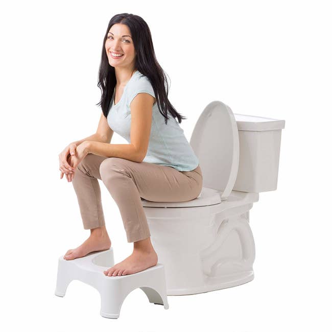Fully clothed model sits on toilet with their feet on the stool, raising their knees above their hips