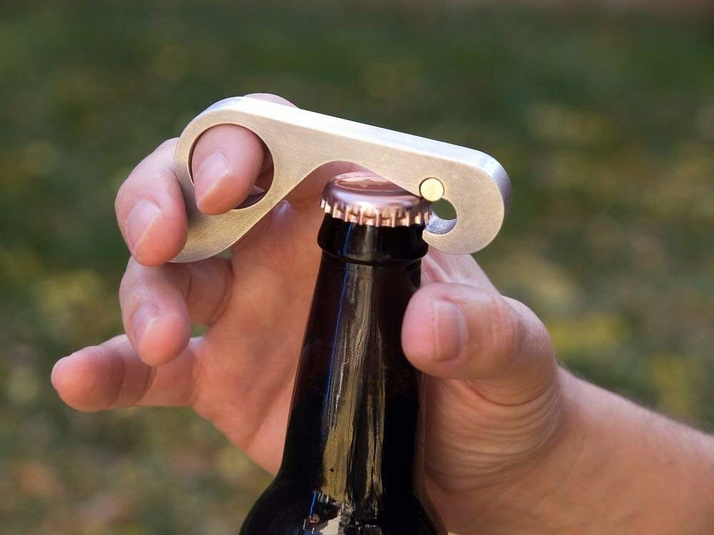 One hand opening a beer with the gold tool, with a finger hole for ease of use