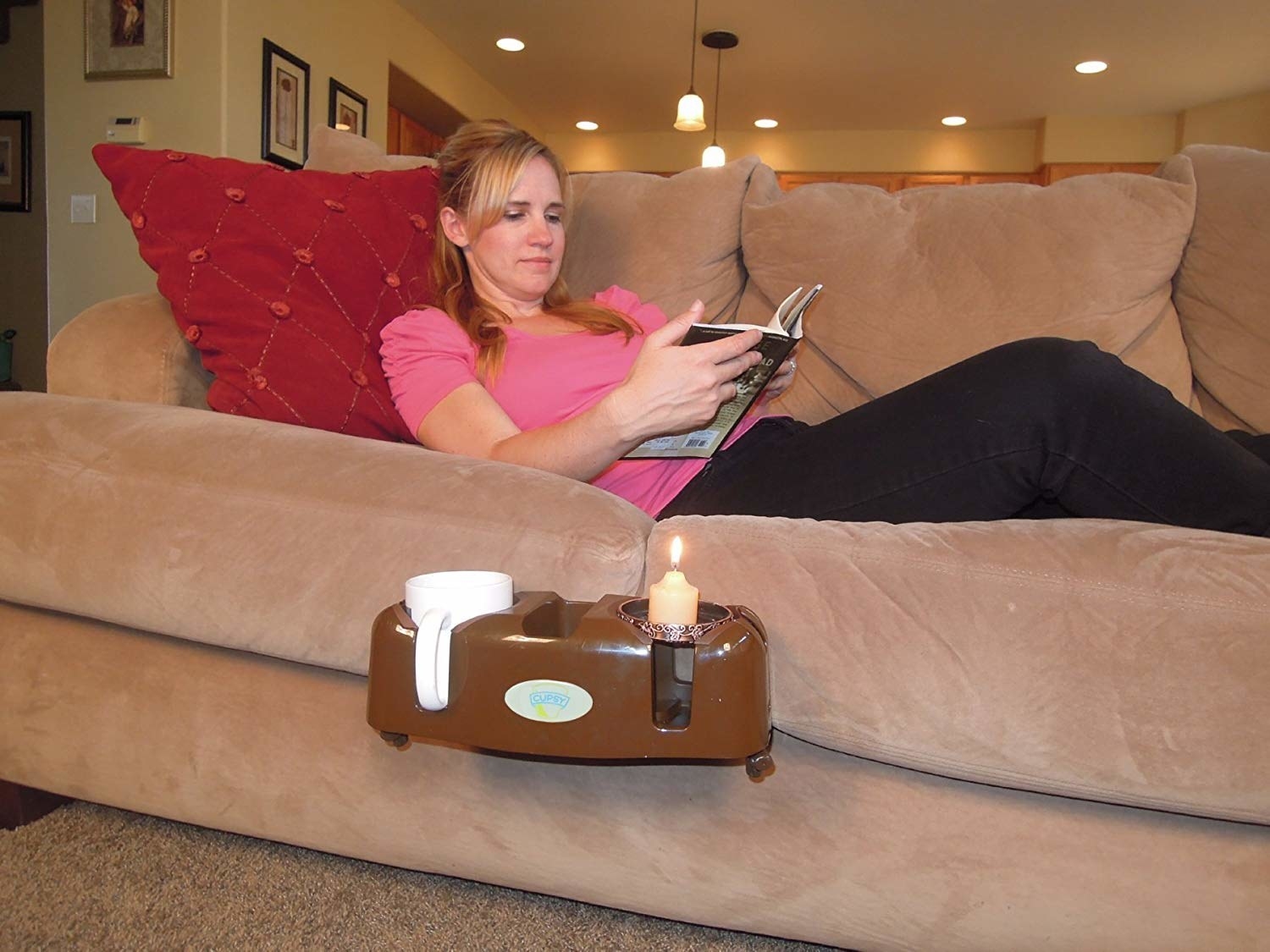 The oval-shaped couch caddy in brown with two cup holders and a smaller holder in the center attached to the side of a couch with a model lying and reading above it