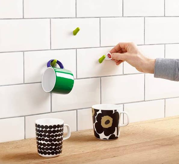 person putting a hook with the moldable glue on a subway tile wall to hang up coffee mugs