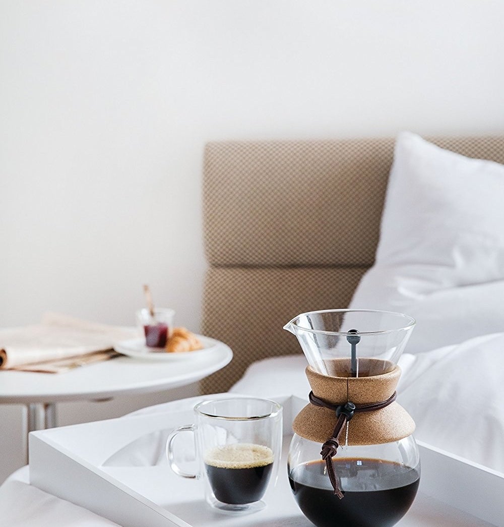the coffee maker on a serving tray next to a cup of coffee placed on a bed with white sheets