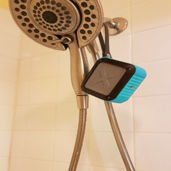 The blue, black, and gray square speaker hanging by the strap from a showerhead
