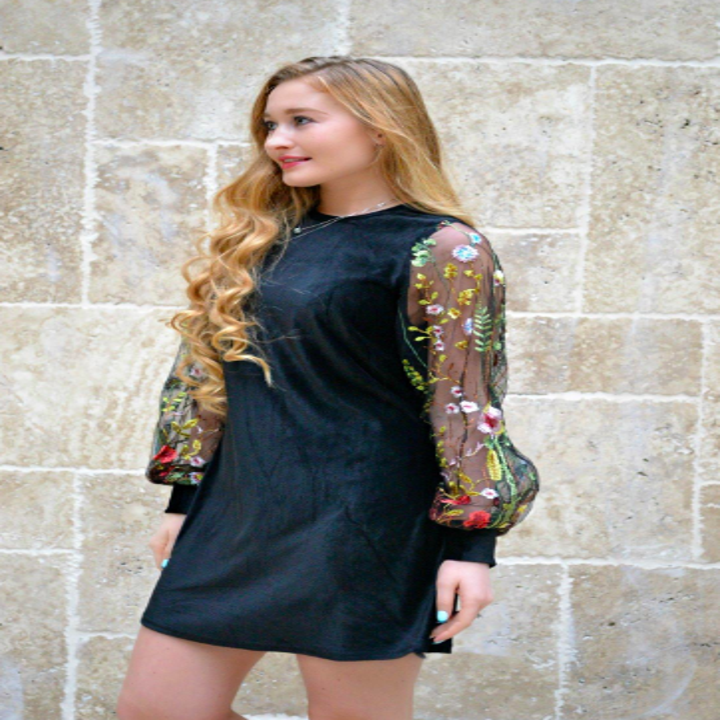A reviewer wearing the dress in black velvet with long sheer sleeves embroidered with colorful flowers