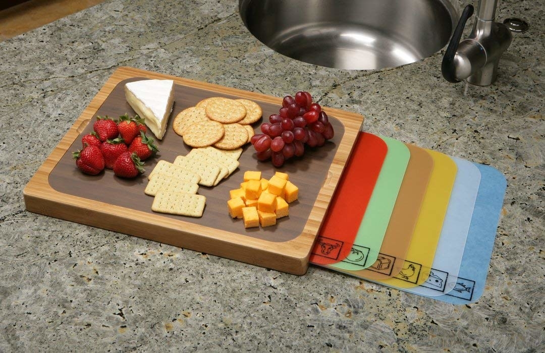 wood cutting board on kitchen counter with gray plastic mat on top of the wood and a cheese board display on top. Six similar plastic mats with icons for types of food are fanned out from beneath the cutting board