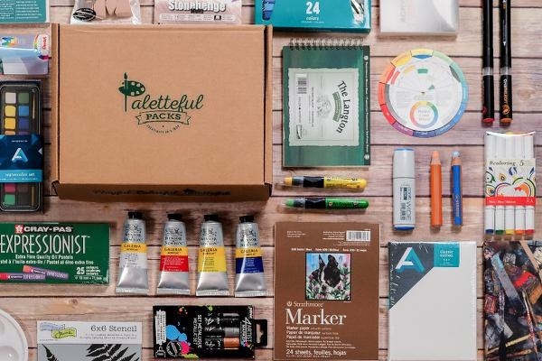 paints, markers, oil pastels, sketchbooks, and other art supplies next to a subscription box