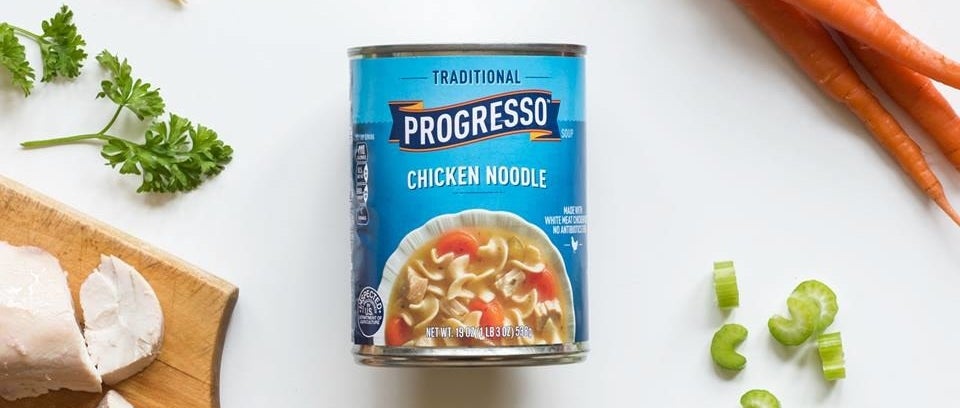 A blue can of Progresso chicken noodle sitting on a wite surface along with chopped celery, carrots, and chicken.