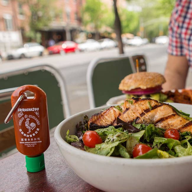 A filled Sriracha bottle keychain beside a meal at a restaurant 