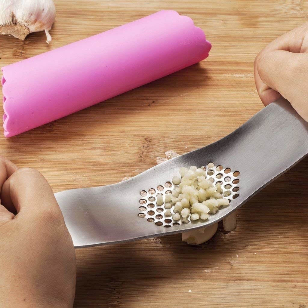 hands holding the silver stainless steel garlic press, mincing some garlic through the wholes in the middle, with the pink round peeler sitting next to it