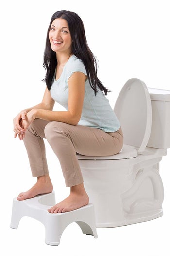 A model sitting on a toilet with their feet propped up on the white Squatty Potty 