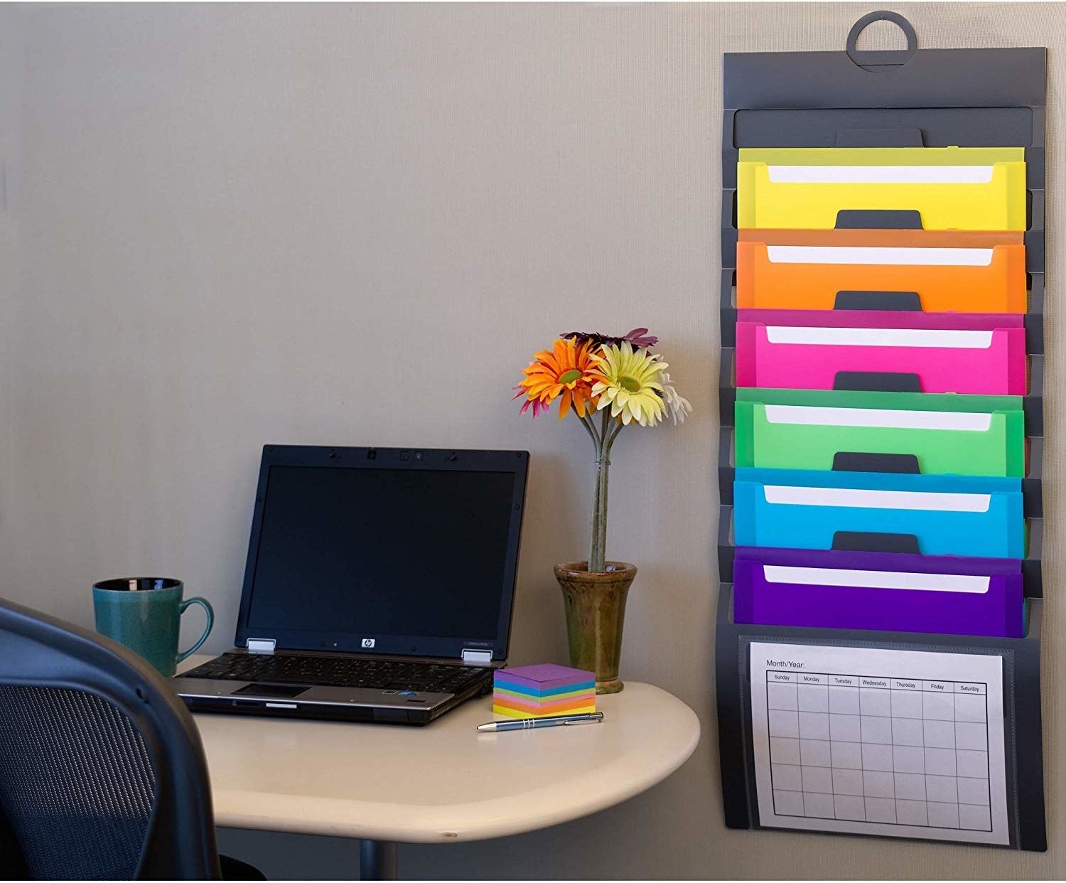 The organizer next to a desk. It lies flat against the wall, with yellow, orange, pink, green, blue, and purple pockets in addition to the clear one