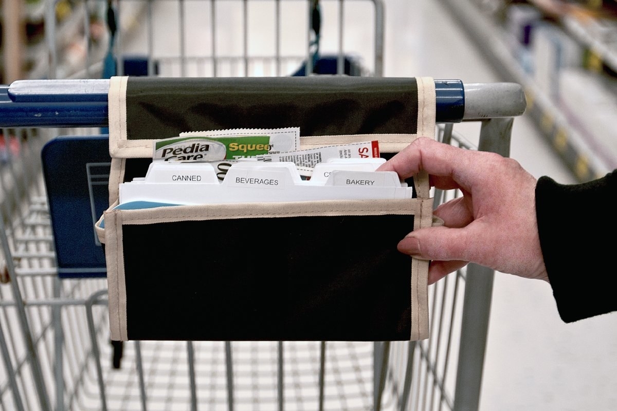 A model&#x27;s hand holds the small organizer, which Velcros over a cart handle and has several labeled dividers like &quot;bakery&quot;, &quot;beverages&quot;, and &quot;canned&quot; 