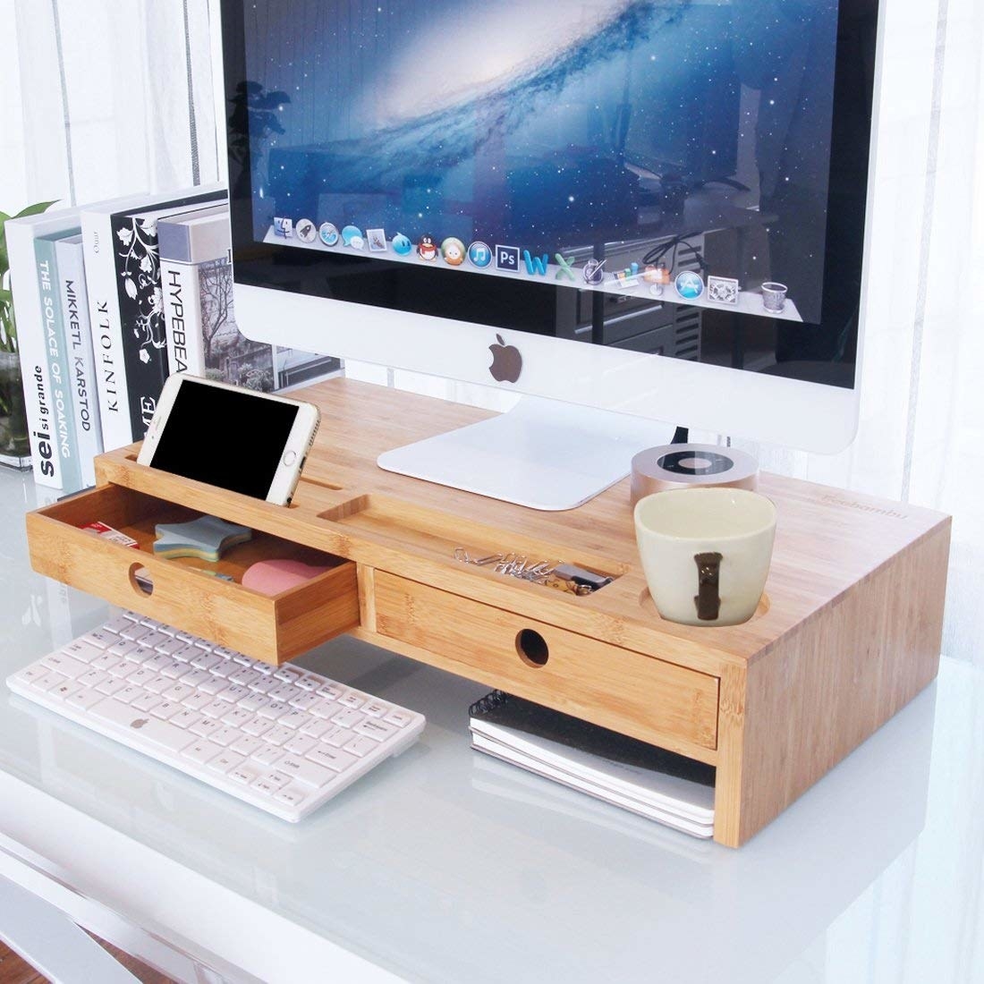 the desktop stand is sitting on a desk. It looks like a mini platform and is made of wood. It has two drawers and space underneath to slide a keyboard under and a notebook.