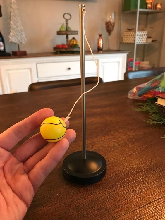 Mini tetherball set on a wooden table with the ball held in someone&#x27;s hand. 