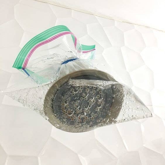 A shower head covered by a sandwich bag filled with vinegar, baking soda, and decalcifier 