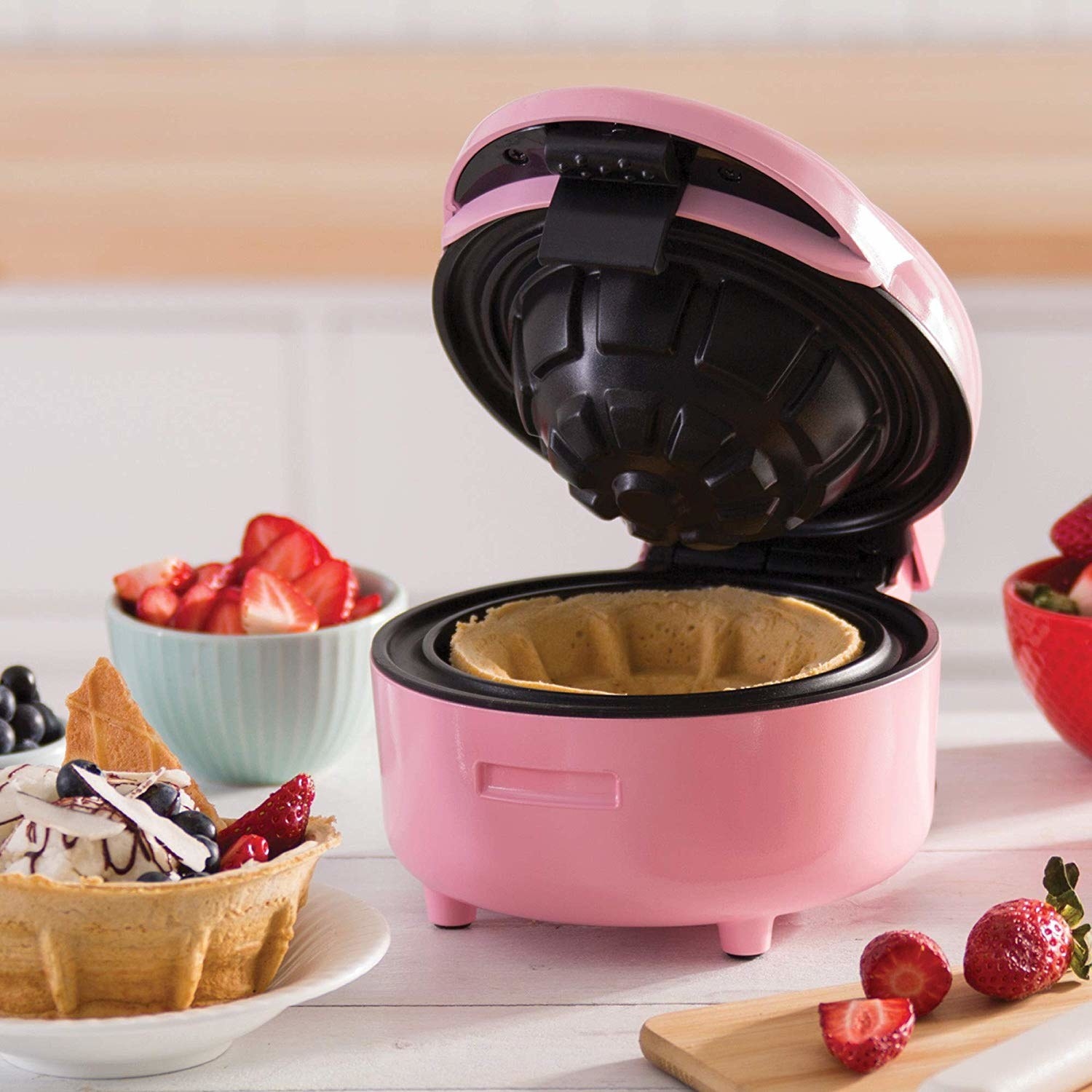 The waffle bowl maker making a waffle bowl, with a made one filled with ice cream, fruit, and more sitting nearby