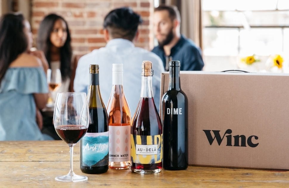 A glasses red wine, four bottles of wine, and a cardboard &quot;Winc&quot; box sit on a wooden table.