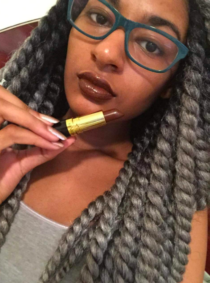 A reviewer wearing a maroon shade and holding up the black and gold tube. The shade in the tube matches the way it looks on their lips