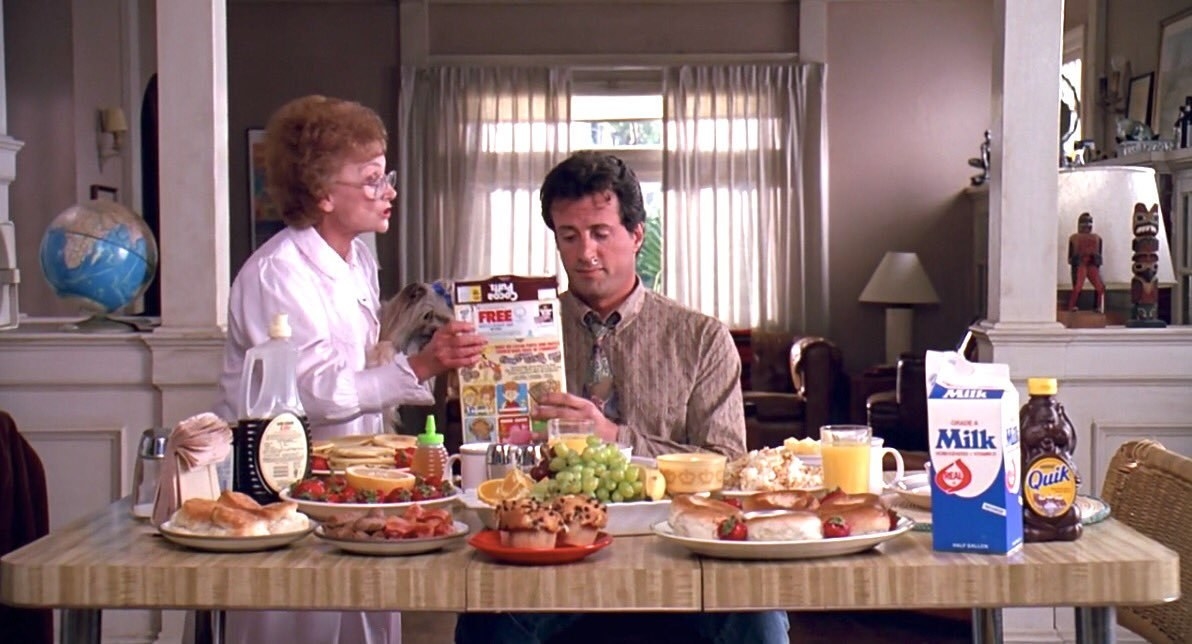 Sylvester Stallone sits at a kitchen table covered in food as Estelle Getty holds a cereal box