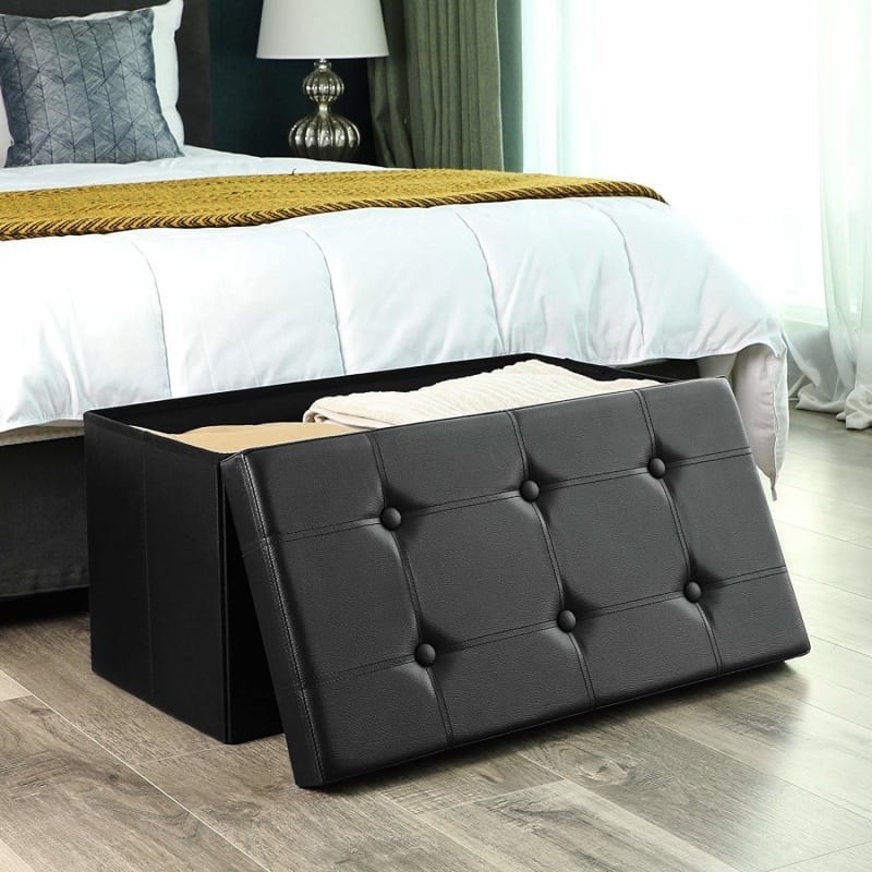 The ottoman sitting in front of a bed with the top leaning against it, showing that it&#x27;s filled with blankets