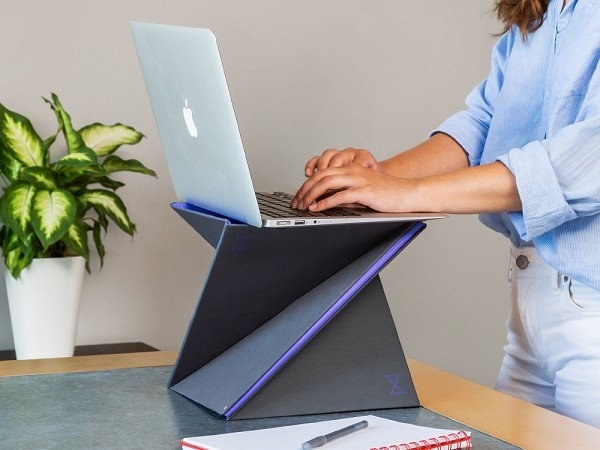 The foldable laptop stand in grey on a table with a laptop on top of it