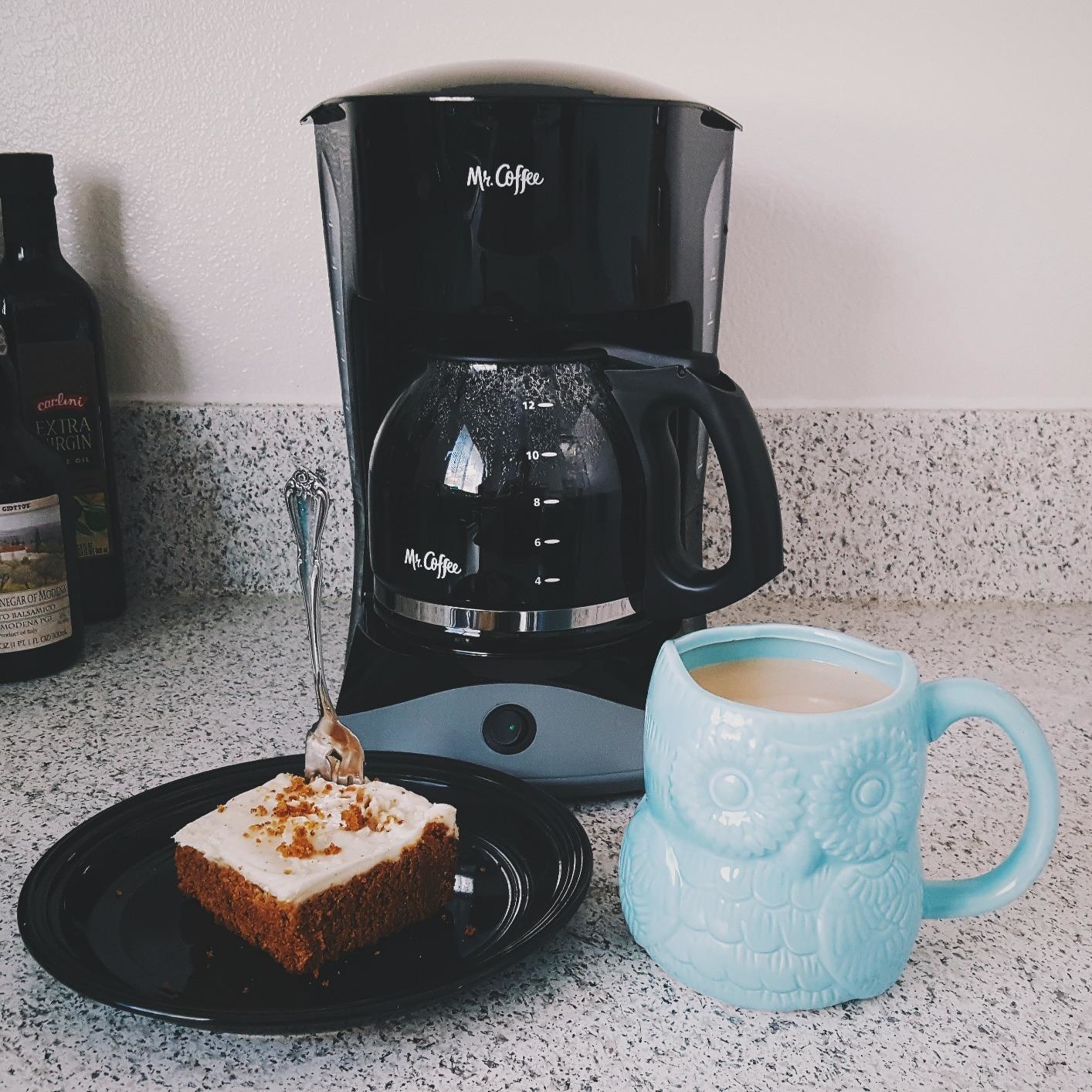 A reviewer showing the coffee maker with an attached kettle and a cup of coffee and a cake