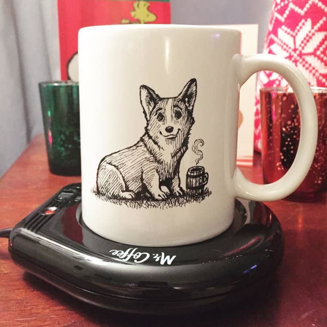 a mug with a drawing of a corgi with a coffee mug on it sitting on a flat black Mr. Coffee mug warmer