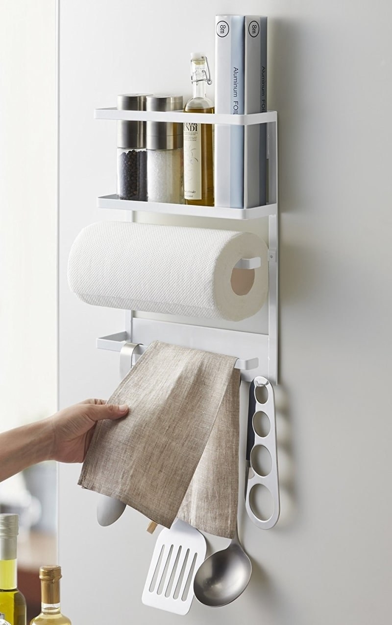A white rack with a shelf with assorted thing such as salt and pepper and oil, then a rod for a paper towel roll, and then another rod for assorted hanging tools and a kitchen towel