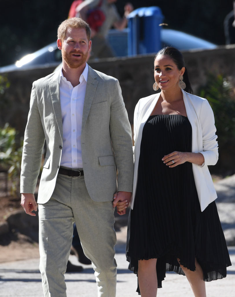 Meghan wears a pleated knee-length dress with a blazer on top alongside Harry wearing a linen suit